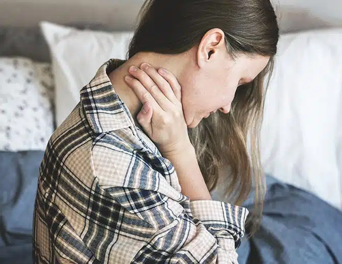 Young woman suffering from neck pain holds her neck while sitting on her bed